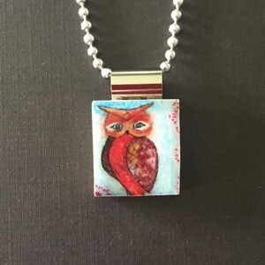 May include: A silver chain necklace with a square pendant featuring a hand-painted owl with red, orange, and yellow feathers on a blue background with white dots.