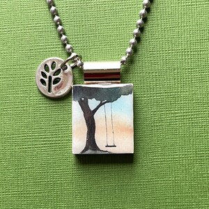 May include: Silver-toned necklace with a square pendant featuring a watercolor painting of a tree and swing. A small, round charm with a tree design hangs next to the pendant. The necklace has a ball chain.