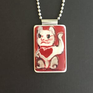 May include: A silver necklace with a rectangular pendant featuring a white cat with a red heart on a red background. The cat has a whimsical design. The pendant is framed in silver and hangs from a silver ball chain.