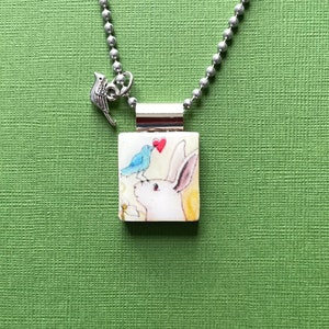 May include: A silver chain necklace with a small bird charm and a square pendant featuring a watercolor illustration of a white rabbit with a blue bird perched on its head. The bird is holding a small red heart.