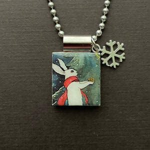 May include: A silver chain necklace with a square pendant featuring a white rabbit wearing a red scarf and holding a small golden ball. A silver snowflake charm hangs from the pendant.
