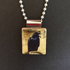 May include: A silver chain necklace with a rectangular pendant featuring a black raven on a yellow background.