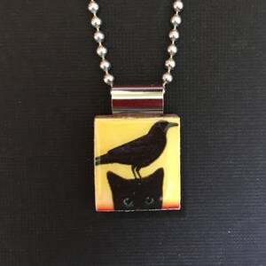 May include: A silver chain necklace with a rectangular pendant featuring a black bird perched on the head of a black cat. The background of the pendant is yellow.