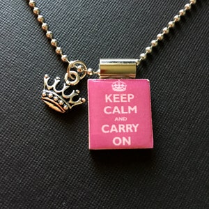 May include: A silver chain necklace with a pink rectangular charm that says "Keep Calm and Carry On". The charm has a silver crown on top and a silver crown on the bottom.