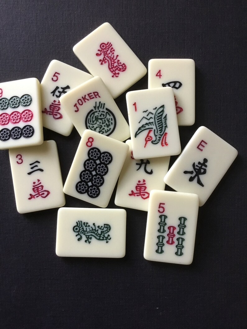 May include: A collection of off-white Mahjong tiles with red, green, and black symbols and characters. Some tiles display numbers, floral patterns, and the word "JOKER". The tiles are scattered on a dark surface.