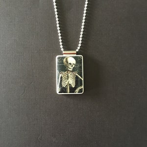 May include: A silver chain necklace with a rectangular pendant featuring a black and white image of a skeleton.