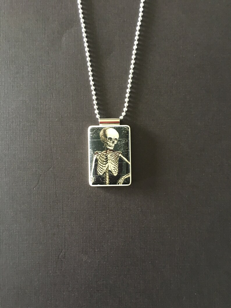 May include: A silver chain necklace with a rectangular pendant featuring a black and white image of a skeleton.