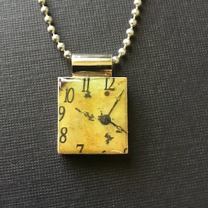 May include: A silver chain necklace with a rectangular pendant featuring a vintage clock face with Roman numerals and black hands. The clock face is yellowed and has a distressed look.