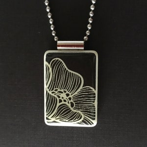 May include: A rectangular pendant necklace with a black background and a white floral design. The pendant is attached to a silver ball chain. The necklace is displayed against a dark background.