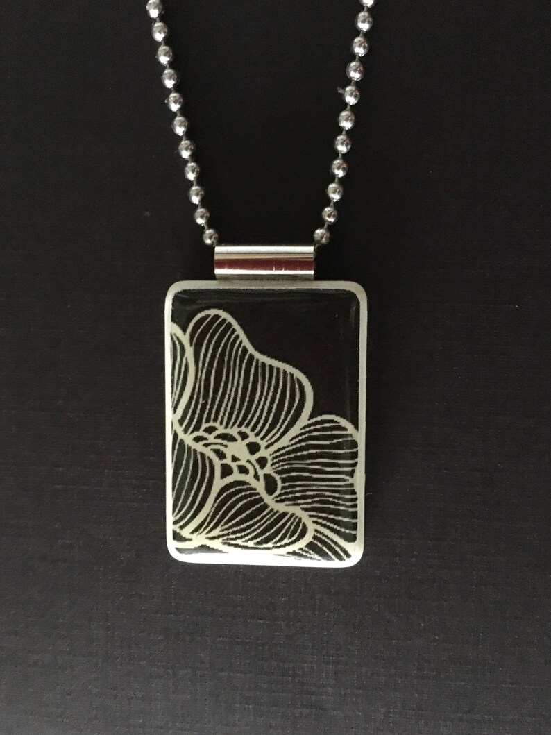 May include: A rectangular pendant necklace with a black background and a white floral design. The pendant is attached to a silver ball chain. The necklace is displayed against a dark background.