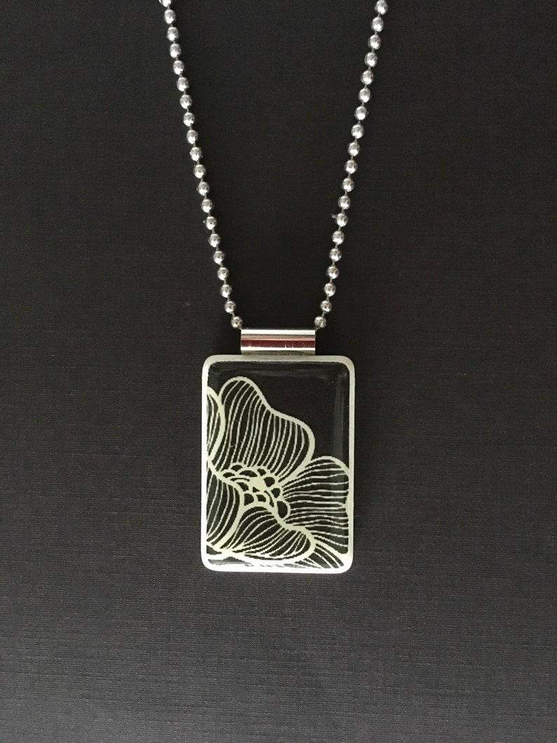 May include: A silver-toned necklace with a rectangular pendant featuring a white floral design on a black background. The pendant is suspended from a ball chain. The necklace is displayed against a dark background.
