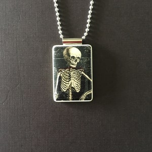 May include: A silver chain necklace with a rectangular pendant featuring a black and white image of a skeleton.