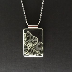 May include: A rectangular pendant necklace with a black background and a white floral design. The pendant is attached to a silver ball chain. The floral design is a detailed line drawing.
