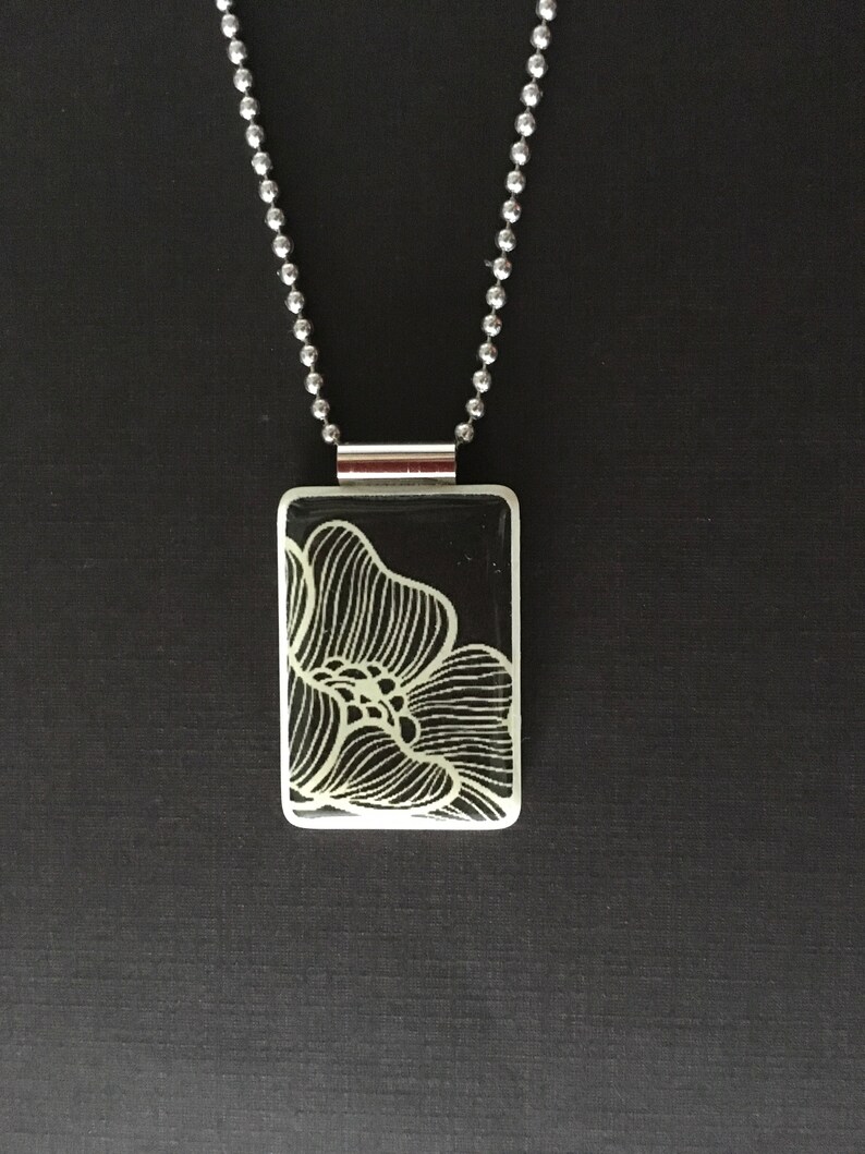 May include: A rectangular pendant necklace with a black background and a white floral design. The pendant is attached to a silver ball chain. The floral design is a detailed line drawing.