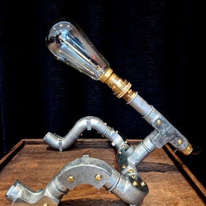 Hand-built industrial robot lamp made from silver galvanised steel conduit on a wooden desk with a glowing Edison bulb.