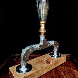 Handcrafted industrial-style table lamp made from galvanized metal pipes and brass fittings, mounted on a solid wooden base with a natural wood grain finish. Its minimalist, steampunk-inspired design adds a rustic and modern touch to any home