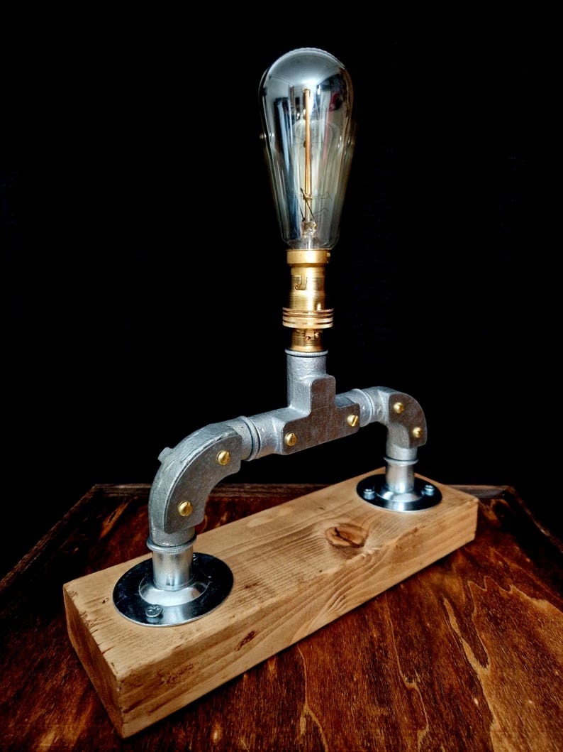 Handcrafted industrial-style table lamp made from galvanized metal pipes and brass fittings, mounted on a solid wooden base with a natural wood grain finish. Its minimalist, steampunk-inspired design adds a rustic and modern touch to any home