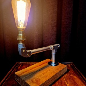 Handcrafted industrial-style table lamp made from galvanized metal pipes and brass fittings, mounted on a solid wooden base with a natural wood grain finish. Its minimalist, steampunk-inspired design adds a rustic and modern touch to any home