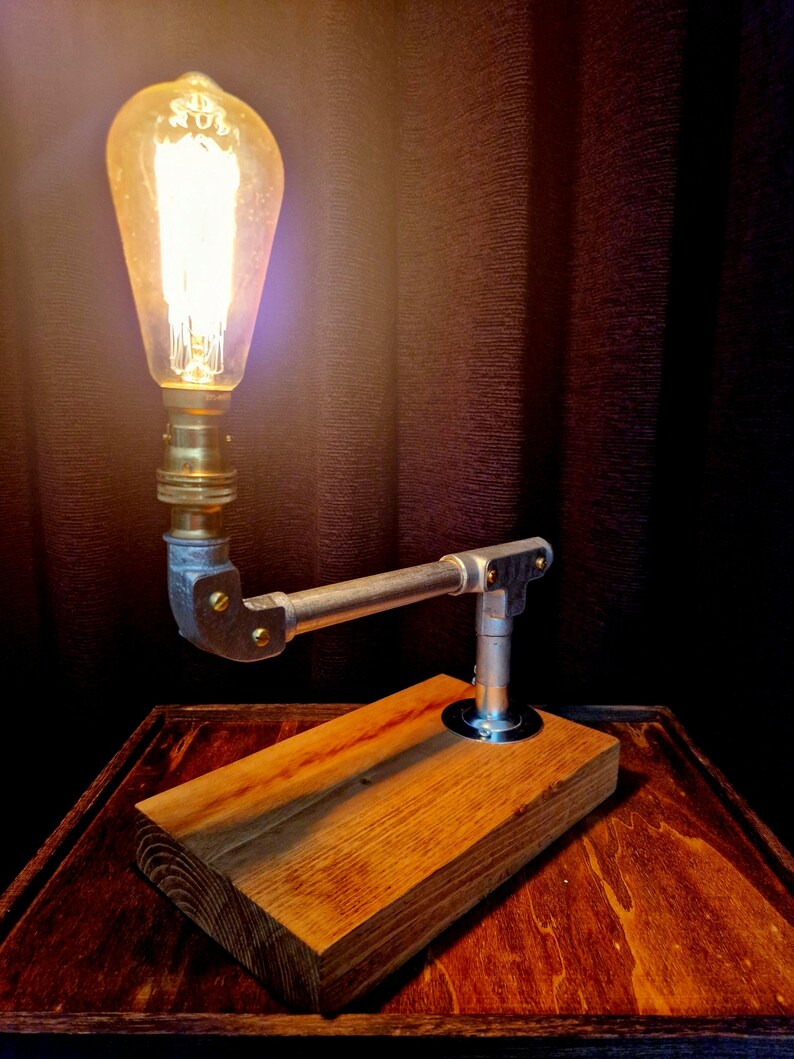 Handcrafted industrial-style table lamp made from galvanized metal pipes and brass fittings, mounted on a solid wooden base with a natural wood grain finish. Its minimalist, steampunk-inspired design adds a rustic and modern touch to any home