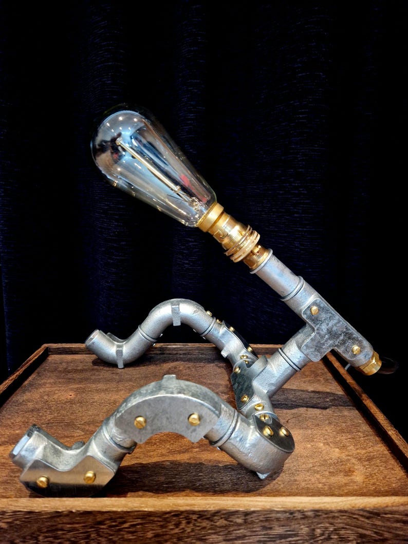 Hand-built industrial robot lamp made from silver galvanised steel conduit on a wooden desk with a glowing Edison bulb.