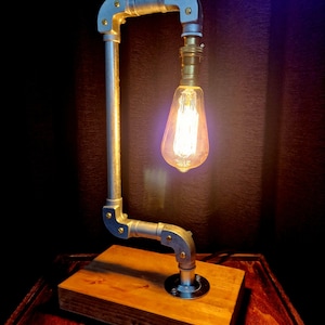May include: A silver pipe table lamp with a wooden base and a vintage-style light bulb. The lamp is switched on and the light bulb is glowing.