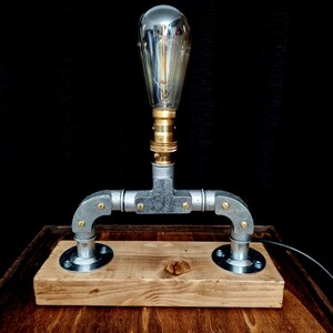Handcrafted industrial-style table lamp made from galvanized metal pipes and brass fittings, mounted on a solid wooden base with a natural wood grain finish. Its minimalist, steampunk-inspired design adds a rustic and modern touch to any home