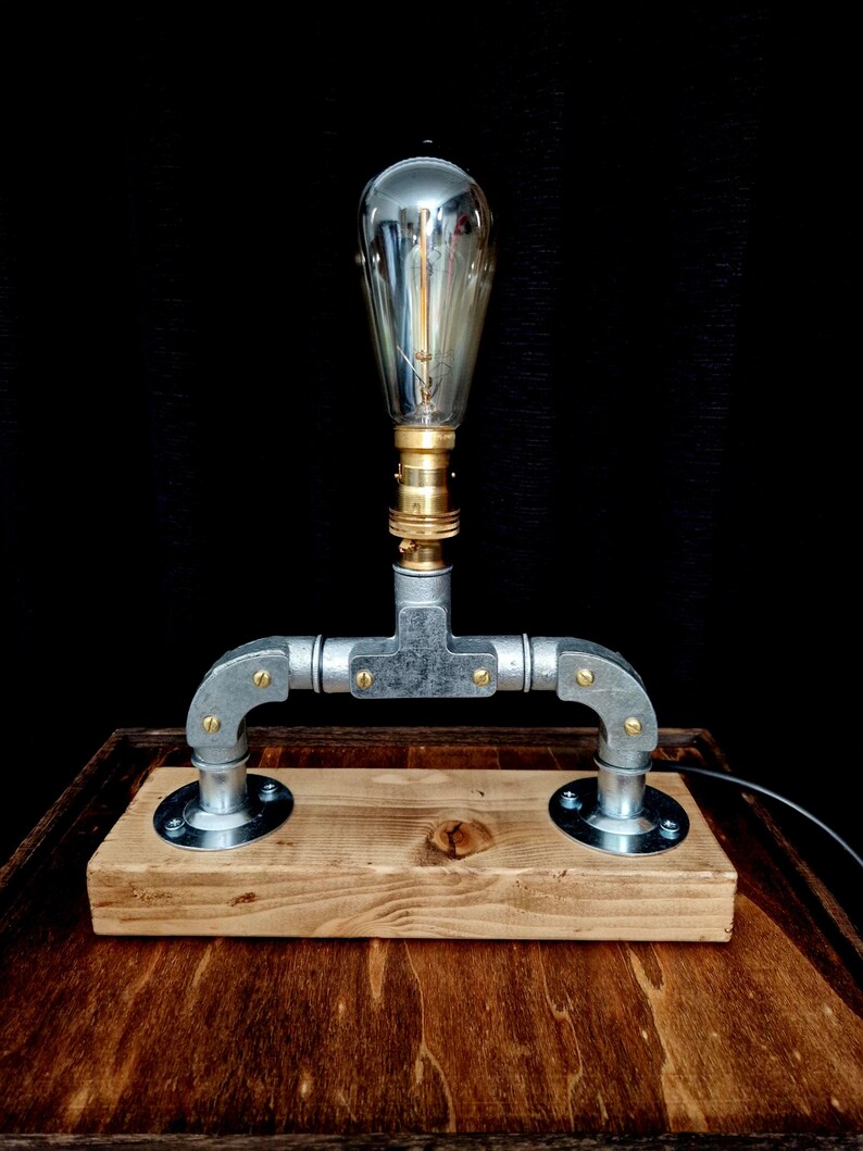 Handcrafted industrial-style table lamp made from galvanized metal pipes and brass fittings, mounted on a solid wooden base with a natural wood grain finish. Its minimalist, steampunk-inspired design adds a rustic and modern touch to any home