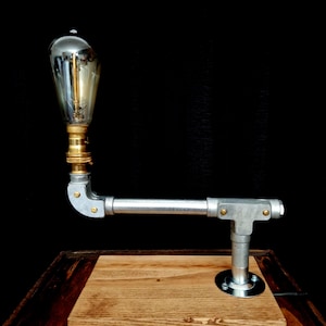 Handcrafted industrial-style table lamp made from galvanized metal pipes and brass fittings, mounted on a solid wooden base with a natural wood grain finish. Its minimalist, steampunk-inspired design adds a rustic and modern touch to any home