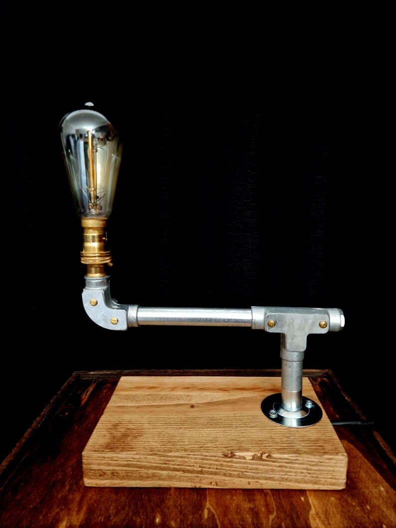 Handcrafted industrial-style table lamp made from galvanized metal pipes and brass fittings, mounted on a solid wooden base with a natural wood grain finish. Its minimalist, steampunk-inspired design adds a rustic and modern touch to any home