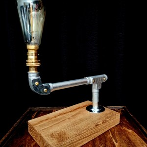 Handcrafted industrial-style table lamp made from galvanized metal pipes and brass fittings, mounted on a solid wooden base with a natural wood grain finish. Its minimalist, steampunk-inspired design adds a rustic and modern touch to any home