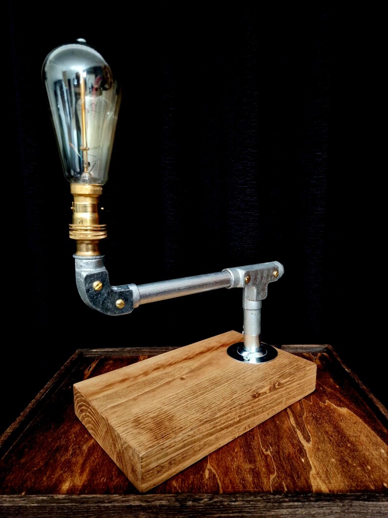 Handcrafted industrial-style table lamp made from galvanized metal pipes and brass fittings, mounted on a solid wooden base with a natural wood grain finish. Its minimalist, steampunk-inspired design adds a rustic and modern touch to any home