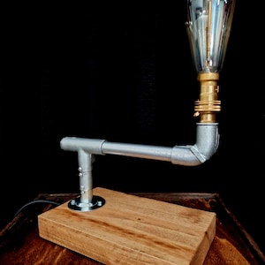 Handcrafted industrial-style table lamp made from galvanized metal pipes and brass fittings, mounted on a solid wooden base with a natural wood grain finish. Its minimalist, steampunk-inspired design adds a rustic and modern touch to any home