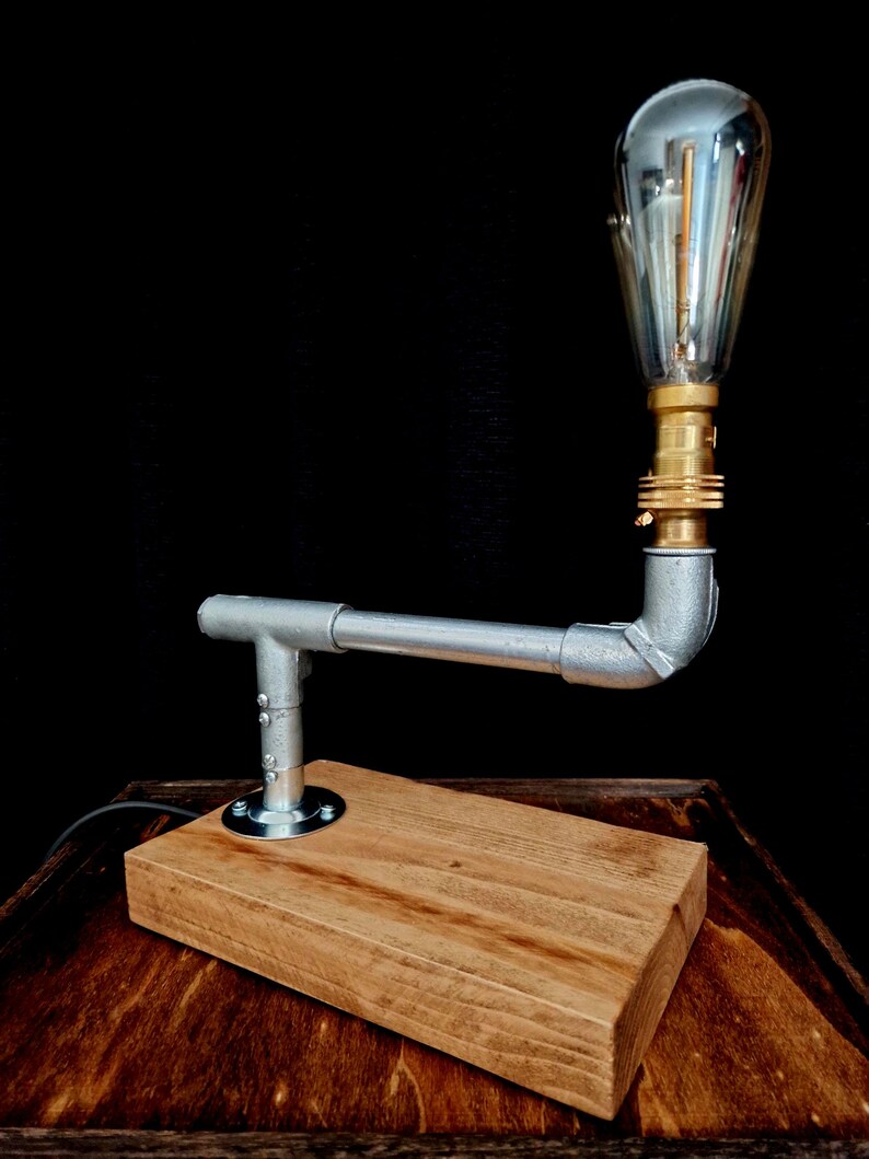 Handcrafted industrial-style table lamp made from galvanized metal pipes and brass fittings, mounted on a solid wooden base with a natural wood grain finish. Its minimalist, steampunk-inspired design adds a rustic and modern touch to any home