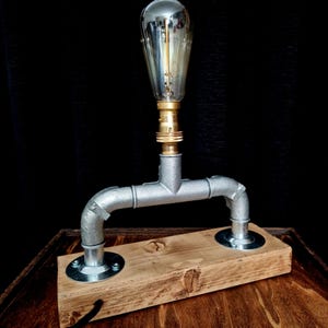 Handcrafted industrial-style table lamp made from galvanized metal pipes and brass fittings, mounted on a solid wooden base with a natural wood grain finish. Its minimalist, steampunk-inspired design adds a rustic and modern touch to any home