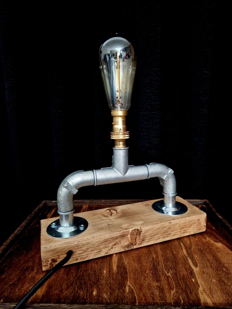 Handcrafted industrial-style table lamp made from galvanized metal pipes and brass fittings, mounted on a solid wooden base with a natural wood grain finish. Its minimalist, steampunk-inspired design adds a rustic and modern touch to any home