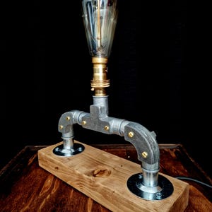 Handcrafted industrial-style table lamp made from galvanized metal pipes and brass fittings, mounted on a solid wooden base with a natural wood grain finish. Its minimalist, steampunk-inspired design adds a rustic and modern touch to any home