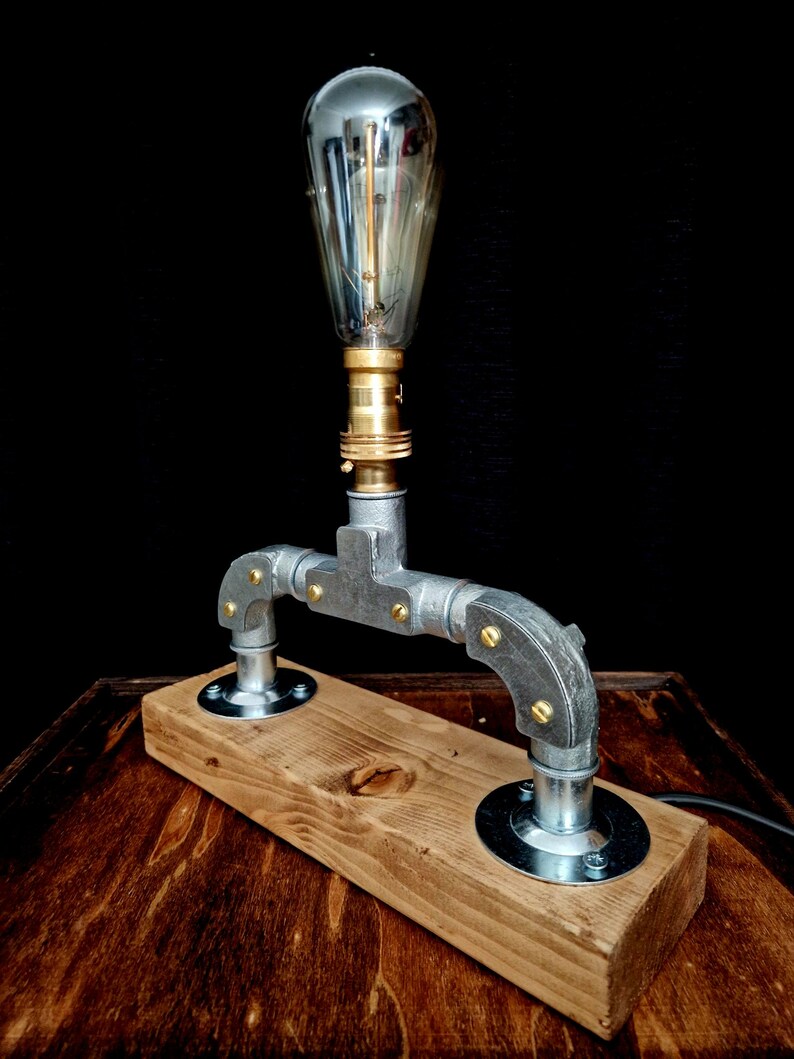 Handcrafted industrial-style table lamp made from galvanized metal pipes and brass fittings, mounted on a solid wooden base with a natural wood grain finish. Its minimalist, steampunk-inspired design adds a rustic and modern touch to any home