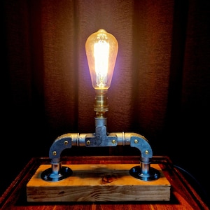 Handcrafted industrial-style table lamp made from galvanized metal pipes and brass fittings, mounted on a solid wooden base with a natural wood grain finish. Its minimalist, steampunk-inspired design adds a rustic and modern touch to any home