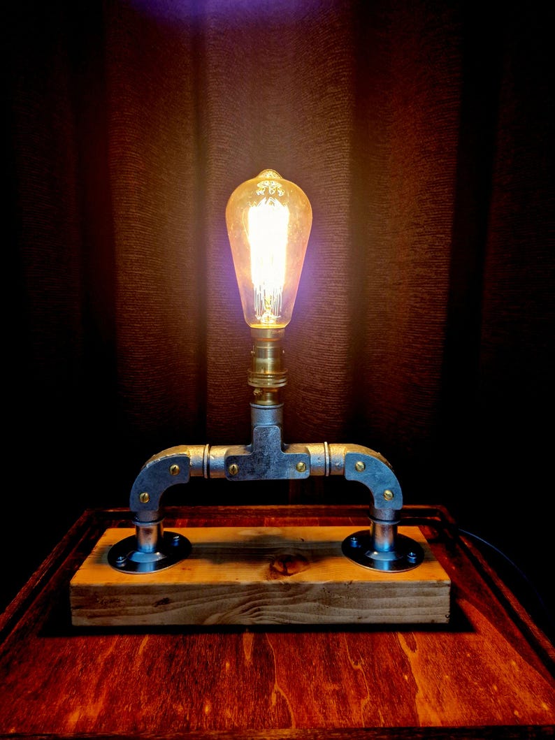 Handcrafted industrial-style table lamp made from galvanized metal pipes and brass fittings, mounted on a solid wooden base with a natural wood grain finish. Its minimalist, steampunk-inspired design adds a rustic and modern touch to any home