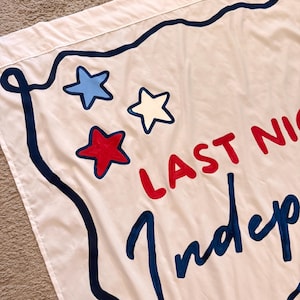 May include: White flag with blue, red, and white stars and text that reads "LAST NIGHT Indep". The flag has a dark blue border and is likely a decorative item.