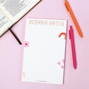 May include: A white notepad with the words "SERMON NOTES" in orange script, decorated with a rainbow, hearts, a flower, and a cross. Two pens, one orange and one pink, are next to the notepad. A Bible is open in the background.