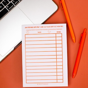 May include: A white task planner with orange text and lines, featuring the months of the year and a space for tasks. Two orange pens and a laptop are on an orange surface. The planner includes the text "SAVVYTHREADS".