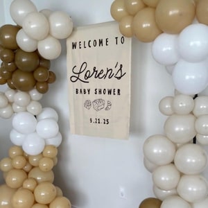 May include: A baby shower decoration featuring balloon arches in white, brown, and tan hues. A banner reads "WELCOME TO Loren's BABY SHOWER" with the date 9.21.25. The balloons are arranged to frame the banner, creating a festive atmosphere.