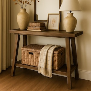 Op de afbeelding: Een houten console met een grote boogvormige spiegel, een vaas met takken, een lamp en een rieten mand. Een ingelijste botanische print en kleine dozen staan op de tafel. Een geruite plaid is over de mand gedrapeerd.