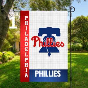 May include: A garden flag featuring the Philadelphia Phillies baseball team's logo. The flag displays a blue Liberty Bell with the word "Phillies" in red script. The word "Philadelphia" is printed vertically in white on a red stripe. The flag is displayed outdoors.