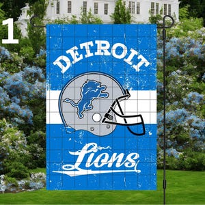 May include: A blue and white Detroit Lions garden flag. The flag features the team's logo, a silver helmet with a blue lion, and the words "DETROIT" and "Lions" in white, distressed lettering. The flag is displayed on a black metal stand.