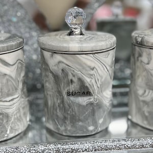 May include: Three grey marble canisters with crystal knobs. The canisters are labelled "Tea", "Sugar", and "Coffee".