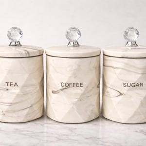 Może przedstawiać: Trzy pasujące ceramiczne pojemniki z pokrywkami, z etykietami "TEA", "COFFEE" i "SUGAR". Pojemniki mają wzór przypominający marmur w kolorze kremowym i szarym, z fasetowanymi bokami. Każda pokrywka jest zwieńczona przezroczystym, kryształowym guzikiem.