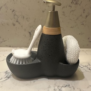 Peut inclure: Un distributeur de savon gris foncé avec une pompe en métal brossé et un accent en bambou. Il comprend une brosse à récurer blanche et une éponge blanche. Le distributeur est posé sur un comptoir, offrant un accessoire de cuisine élégant.