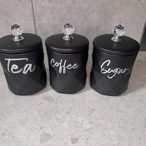 Può includere: Tre barattoli da cucina neri con pomelli a forma di cristallo. I barattoli sono etichettati con "Tea", "Coffee" e "Sugar" in scrittura bianca. I barattoli hanno un design geometrico.